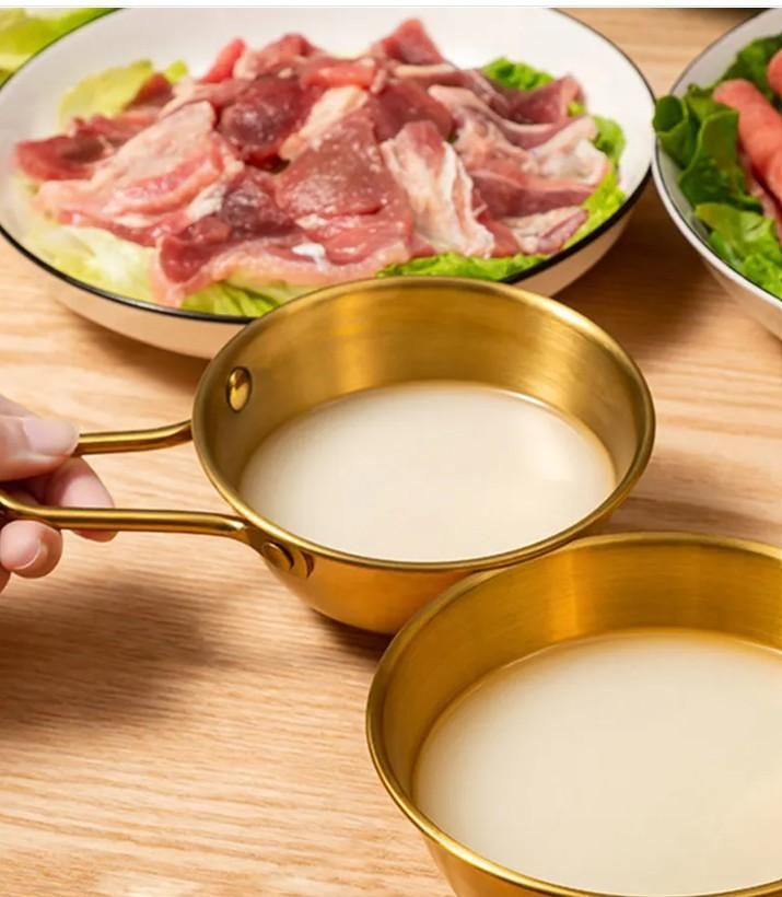 Gold kitchen bowl with handle for ramen containing white broth with plates of raw meat and vegetables on wooden table
